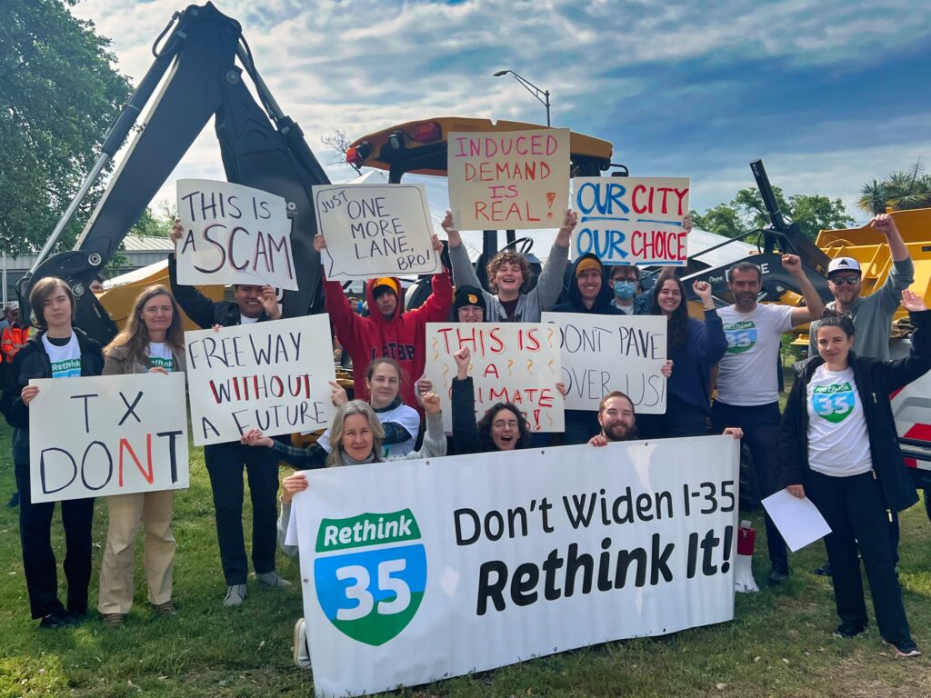 Advocates in Austin in front of bulldozers at I-35 groundbreaking, holding signs protesting the highway expansion. TxDONT; Freeway without a future; This is a scam; Just one more lane bro; induced demand is real; Our city our choice; don't pave over us; this is a climate crisis; Don't widen I-35, Rethink it!